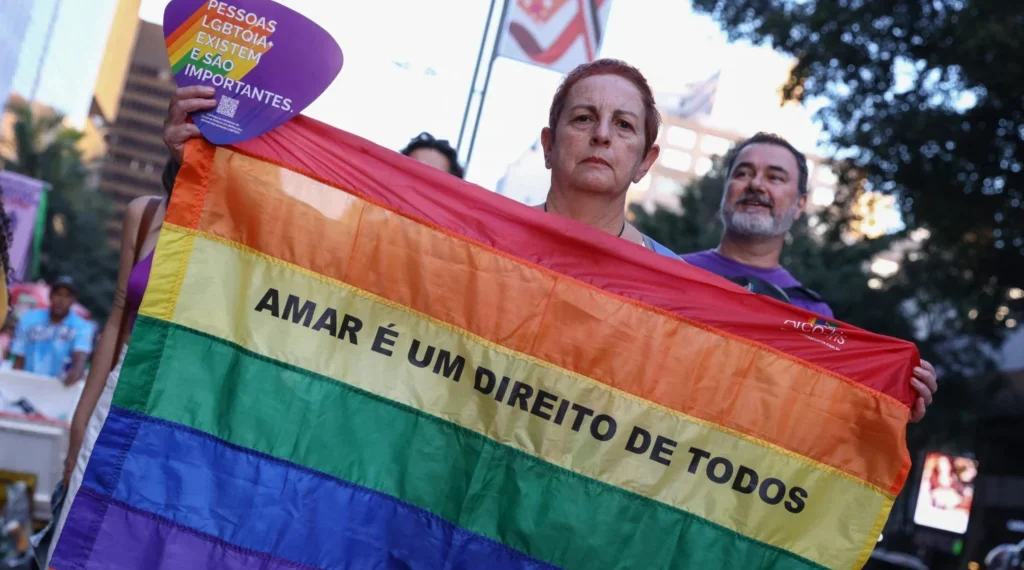 Foto em ambiente urbano. Uma pessoa segura uma bandeira do orgulho LGBTQIAPN+. No centro da bandeira está escrito em letras pretas "Amar é um direito de todos". A pessoa também segura um leque roxo com detalhes do arco-íris e a frase “Pessoas LGBTQIA+ existem e são importantes”
