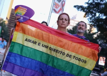 Foto em ambiente urbano. Uma pessoa segura uma bandeira do orgulho LGBTQIAPN+. No centro da bandeira está escrito em letras pretas "Amar é um direito de todos". A pessoa também segura um leque roxo com detalhes do arco-íris e a frase “Pessoas LGBTQIA+ existem e são importantes”
