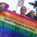 Foto em ambiente urbano. Uma pessoa segura uma bandeira do orgulho LGBTQIAPN+. No centro da bandeira está escrito em letras pretas "Amar é um direito de todos". A pessoa também segura um leque roxo com detalhes do arco-íris e a frase “Pessoas LGBTQIA+ existem e são importantes”